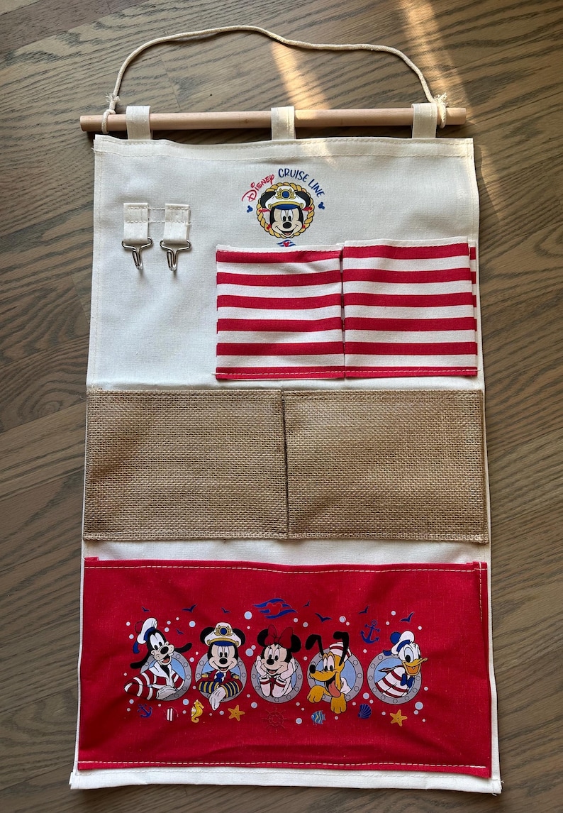 May include: A canvas wall organizer featuring the Disney Cruise Line logo and Mickey Mouse. It has red and white striped pockets, burlap pockets, and a red bottom panel with Disney characters. Includes a wooden dowel and hanging cord.