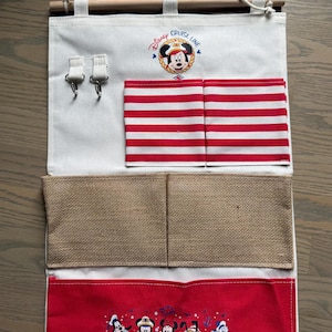 May include: A white fabric organizer with a wooden rod and rope for hanging. It features red and white striped pockets, burlap pockets, and a red bottom panel with Disney characters. The top has the text "Disney Cruise Line" and a Mickey Mouse graphic.