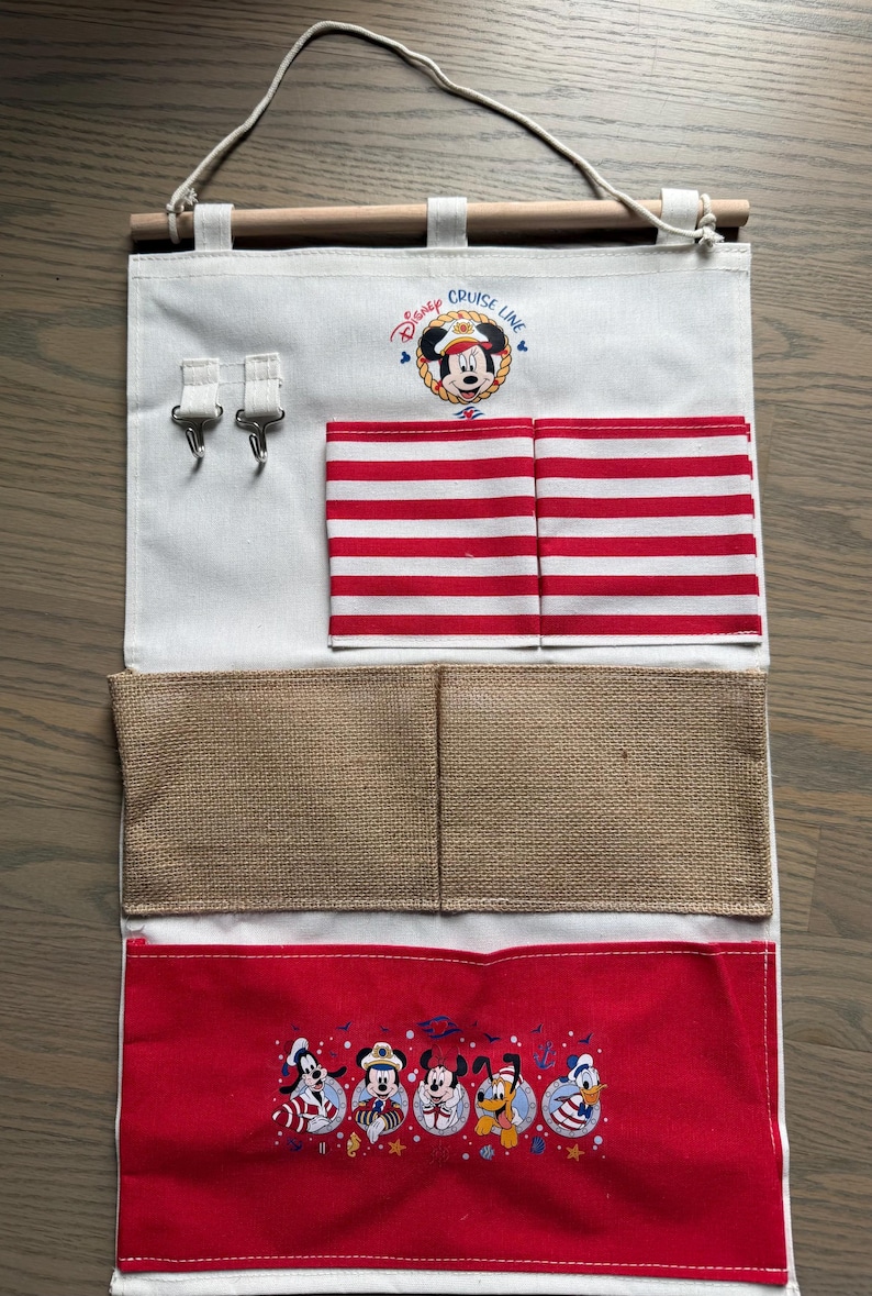 May include: A white fabric organizer with a wooden rod and rope for hanging. It features red and white striped pockets, burlap pockets, and a red bottom panel with Disney characters. The top has the text "Disney Cruise Line" and a Mickey Mouse graphic.