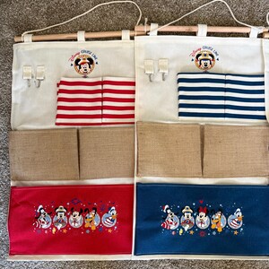 May include: Two hanging organizers with a Disney Cruise Line theme. Each has a wooden rod, cream-colored fabric, and multiple pockets. One has red and white striped pockets, the other has blue and white striped pockets. Both have Mickey Mouse embroidery.