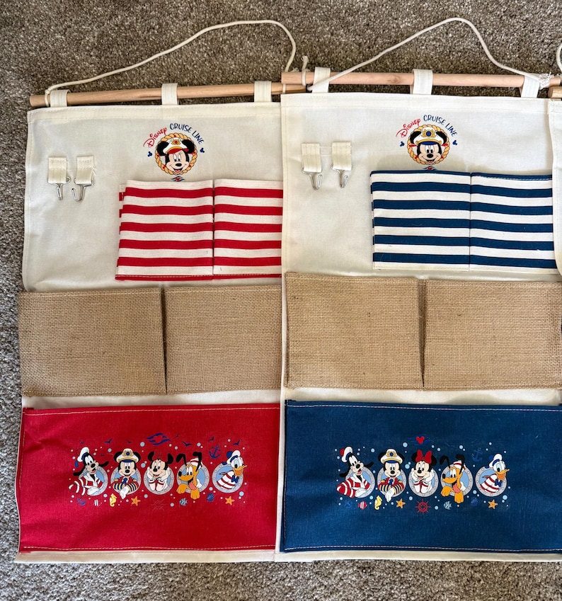 May include: Two hanging organizers with a Disney Cruise Line theme. Each has a wooden rod, cream-colored fabric, and multiple pockets. One has red and white striped pockets, the other has blue and white striped pockets. Both have Mickey Mouse embroidery.