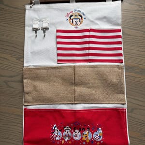 May include: A white fabric organizer with a wooden rod and hanging string. Features red and white striped pockets, burlap pockets, and a red pocket with Disney characters. The top has the text "Disney Cruise Line".