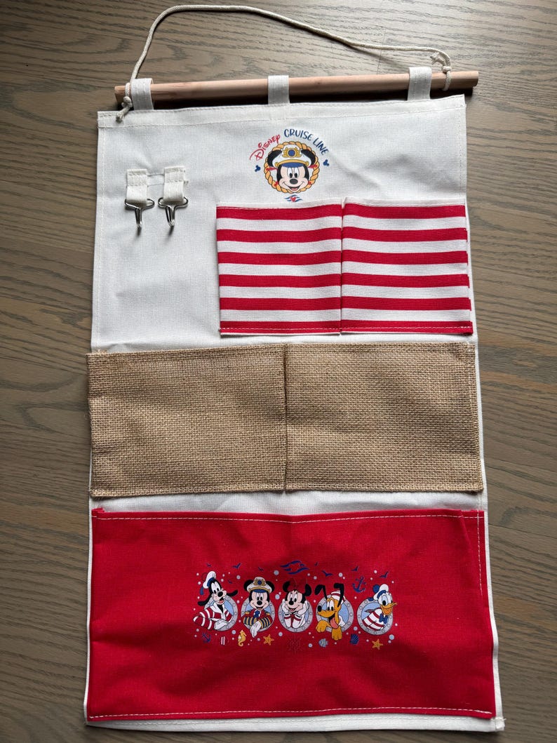 May include: A white fabric organizer with a wooden rod and hanging string. Features red and white striped pockets, burlap pockets, and a red pocket with Disney characters. The top has the text "Disney Cruise Line".