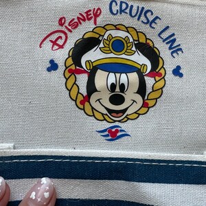 May include: Canvas tote bag featuring a nautical-themed Mickey Mouse graphic. The design includes Mickey wearing a captain's hat, surrounded by a rope border, with "Disney Cruise Line" text. The bag has a blue and white striped pocket.