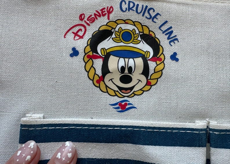 May include: Canvas tote bag featuring a nautical-themed Mickey Mouse graphic. The design includes Mickey wearing a captain's hat, surrounded by a rope border, with "Disney Cruise Line" text. The bag has a blue and white striped pocket.