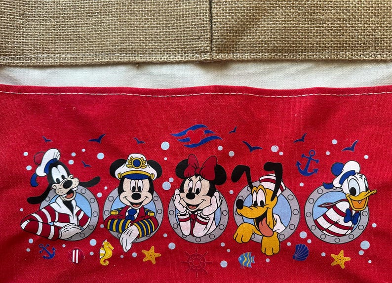 May include: Red fabric featuring Disney characters Goofy, Mickey Mouse, Minnie Mouse, Pluto, and Donald Duck in sailor outfits. The design includes anchors, stars, and other nautical elements, with a burlap and white fabric background.
