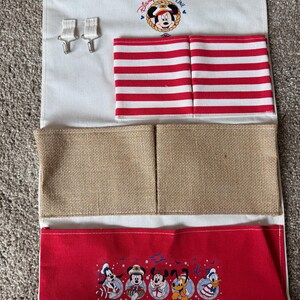 May include: A white fabric organizer with a wooden rod and hooks. It features red and white striped pockets, burlap pockets, and a red bottom section with Disney characters. The top has a Disney Cruise Line logo with Mickey Mouse.