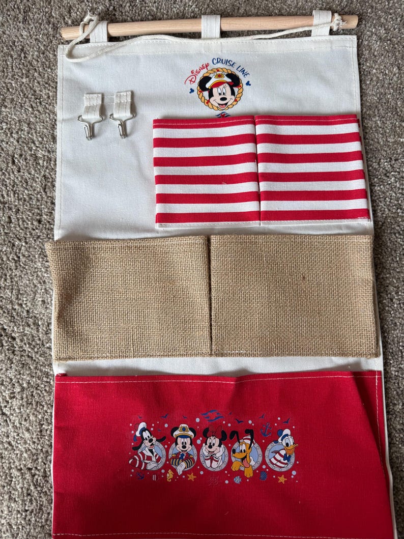 May include: A white fabric organizer with a wooden rod and hooks. It features red and white striped pockets, burlap pockets, and a red bottom section with Disney characters. The top has a Disney Cruise Line logo with Mickey Mouse.