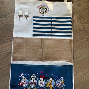 May include: A canvas wall organizer with a wooden dowel and rope for hanging. Features pockets, hooks, and a navy blue bottom panel with Disney Cruise Line characters. Includes Mickey Mouse and friends.