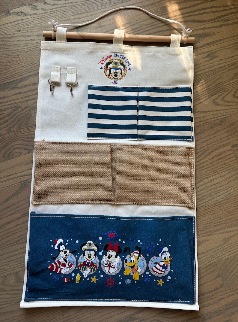 May include: A canvas wall organizer with a wooden dowel and rope for hanging. Features pockets, hooks, and a navy blue bottom panel with Disney Cruise Line characters. Includes Mickey Mouse and friends.