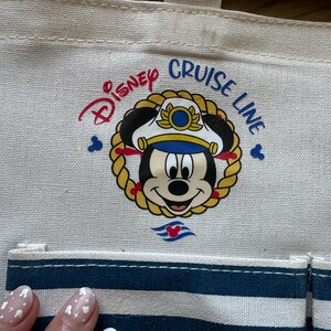 May include: Cream-colored tote bag featuring the Disney Cruise Line logo. The logo includes Mickey Mouse in a captain's hat, surrounded by a gold rope design. The bag has a blue and white striped pocket and the words "Disney Cruise Line" in red and blue.