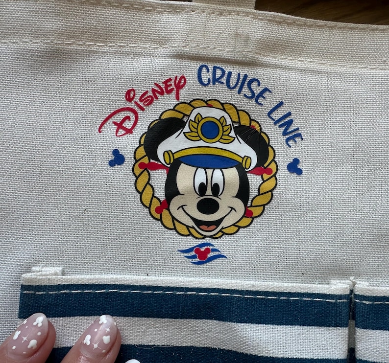 May include: Cream-colored tote bag featuring the Disney Cruise Line logo. The logo includes Mickey Mouse in a captain's hat, surrounded by a gold rope design. The bag has a blue and white striped pocket and the words "Disney Cruise Line" in red and blue.