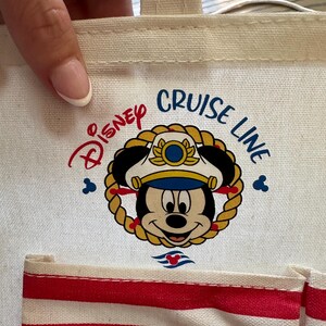 May include: A white canvas tote bag featuring the Disney Cruise Line logo with Mickey Mouse in a captain's hat. The bag has red and white striped pockets and a beige handle.