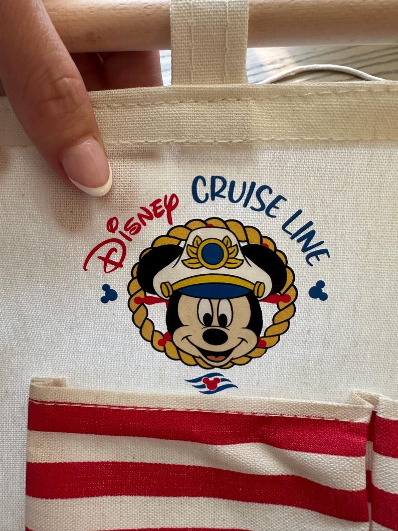May include: A white canvas tote bag featuring the Disney Cruise Line logo with Mickey Mouse in a captain's hat. The bag has red and white striped pockets and a beige handle.