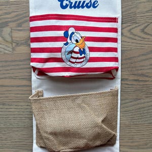 May include: A Disney Cruise hanging organizer with three pockets. The top pocket has the words "Disney Cruise" in blue script. The middle pocket has red and white stripes with a Donald Duck graphic. The bottom pocket is red with a Daisy Duck graphic.