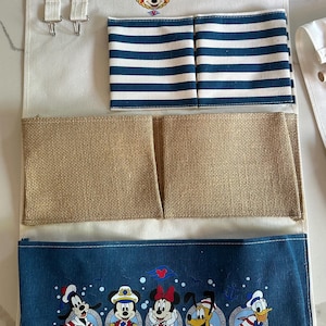 May include: A Disney Cruise Line hanging organizer with multiple pockets. The top section has a wooden rod and hooks. Below are pockets with blue and white stripes, burlap, and a blue fabric with Disney characters.