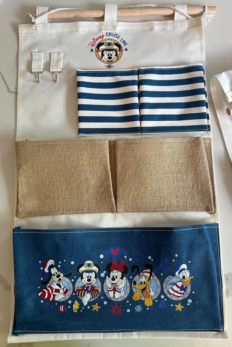 May include: A Disney Cruise Line hanging organizer with multiple pockets. The top section has a wooden rod and hooks. Below are pockets with blue and white stripes, burlap, and a blue fabric with Disney characters.