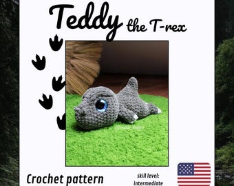 May include: A crocheted gray T-rex dinosaur toy with large blue eyes, lying on green turf. The image includes the text "Teddy the T-rex" and "Crochet pattern" with an American flag. Skill level: intermediate.