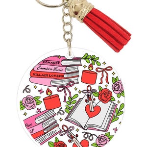 May include: Round keychain with a red tassel. The keychain features a design with stacks of pink books, candles, roses, and an open book with a heart. The books have the titles "Romance", "Enemies to Lovers", and "Villain Lovers".