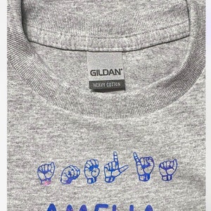 以下が含まれることがあります： クルーネックのグレーのギルダンヘビーコットンTシャツ。シャツには、アメリカ手話で「アメリア」と綴る文字を示す青と紫のデザインが施されています。「ギルダン」の文字がタグに印刷されています。