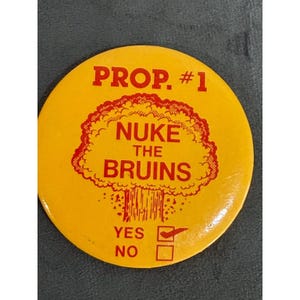 May include: A round yellow pinback button featuring red text that reads PROP. #1 and NUKE THE BRUINS above a graphic of a mushroom cloud. Below the graphic, the text reads YES and NO with a red checkmark inside the box next to the word YES.