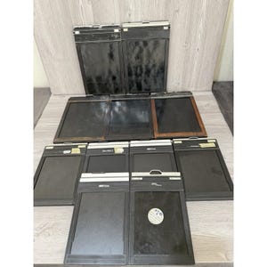 May include: A collection of ten rectangular film holders for large format cameras, arranged in three rows. These black and dark brown holders feature metallic top bars, some with white labels or text, and smooth, dark central panels for light-tight storage.