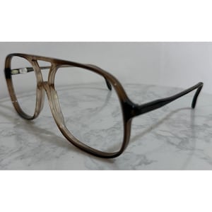 May include: Vintage eyeglasses with a brown and clear frame. The lenses are clear, and the arms are dark brown. The glasses have a double bridge design. The frame is set against a neutral background.