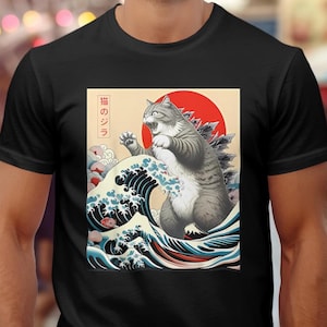 May include: A black t-shirt with a graphic print of a cat in a Godzilla pose, standing on a wave with the Japanese text "猫のジラ" which translates to "Catzilla".