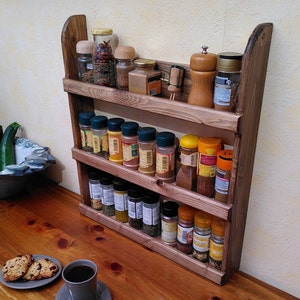 May include: A rustic, three-tiered wooden spice rack filled with various spice jars and containers. The rack is made of stained wood and has a curved top. The spices are arranged on a wooden countertop, with a cup of coffee and cookies in the foreground.