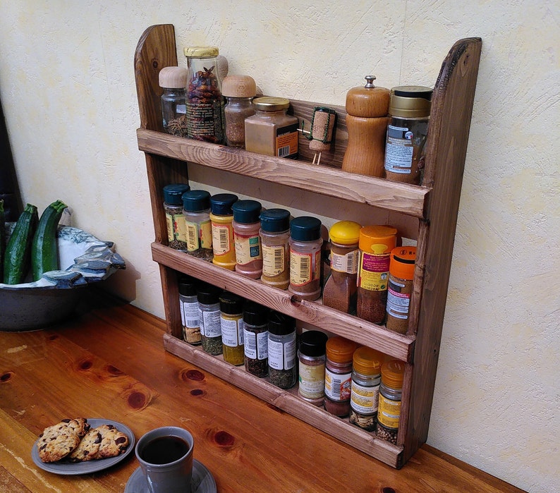 May include: A rustic, three-tiered wooden spice rack filled with various spice jars and containers. The rack is made of stained wood and has a curved top. The spices are arranged on a wooden countertop, with a cup of coffee and cookies in the foreground.