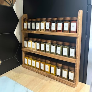 May include: A three-tiered wooden spice rack filled with glass jars of various spices. The jars have wooden lids and white labels. The rack is made of light brown wood and is displayed on a light-colored countertop against a black wall.