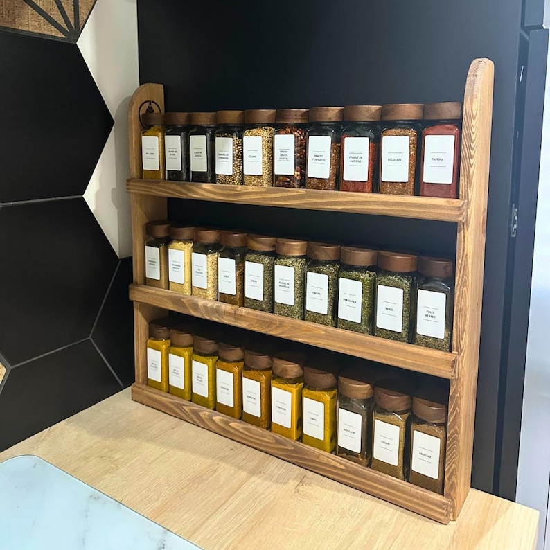 May include: A three-tiered wooden spice rack filled with glass jars of various spices. The jars have wooden lids and white labels. The rack is made of light brown wood and is displayed on a light-colored countertop against a black wall.