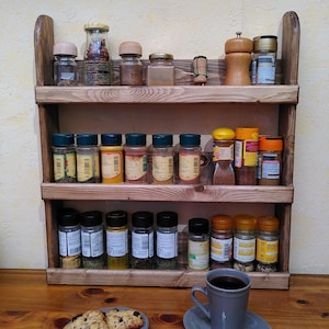 May include: A three-tiered wooden spice rack filled with various spice jars and a pepper grinder. A gray mug of coffee and a plate of chocolate chip cookies sit on the wooden surface in front of the rack.
