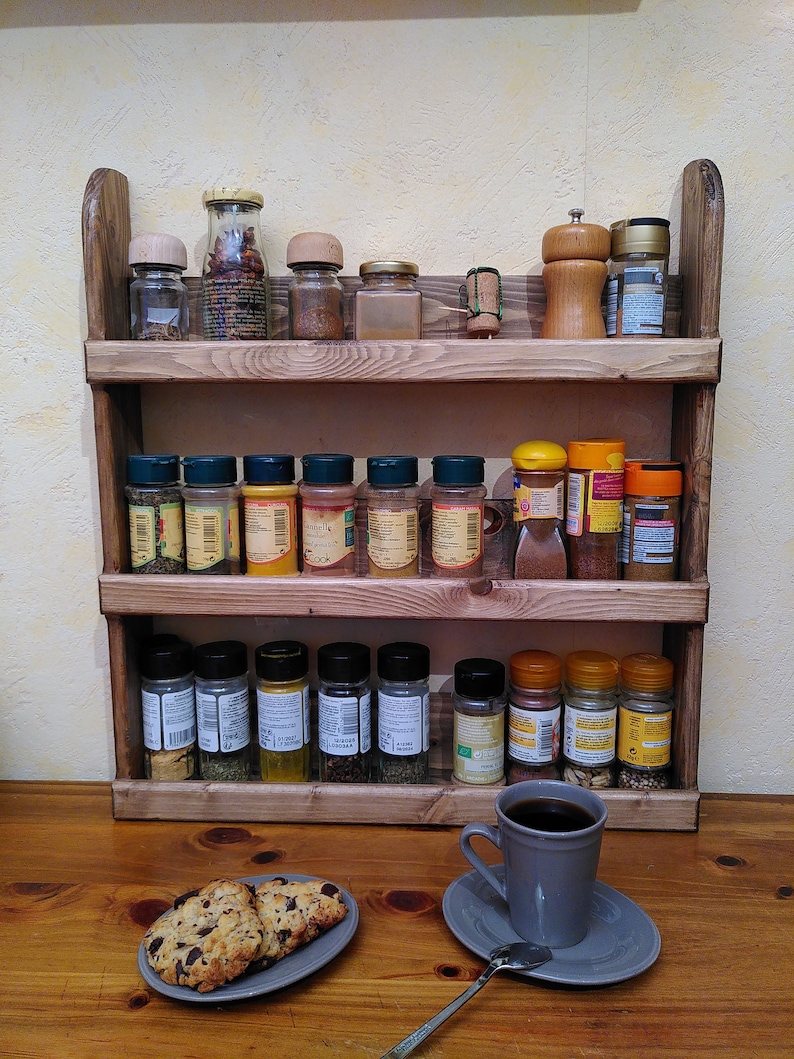 May include: A three-tiered wooden spice rack filled with various spice jars and a pepper grinder. A gray mug of coffee and a plate of chocolate chip cookies sit on the wooden surface in front of the rack.