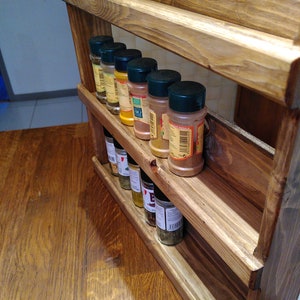 May include: A three-tiered, wooden spice rack with a natural finish. The rack holds several spice jars with black lids, arranged on each shelf. The rack is designed to be mounted on a wall or placed on a countertop for easy access to spices.