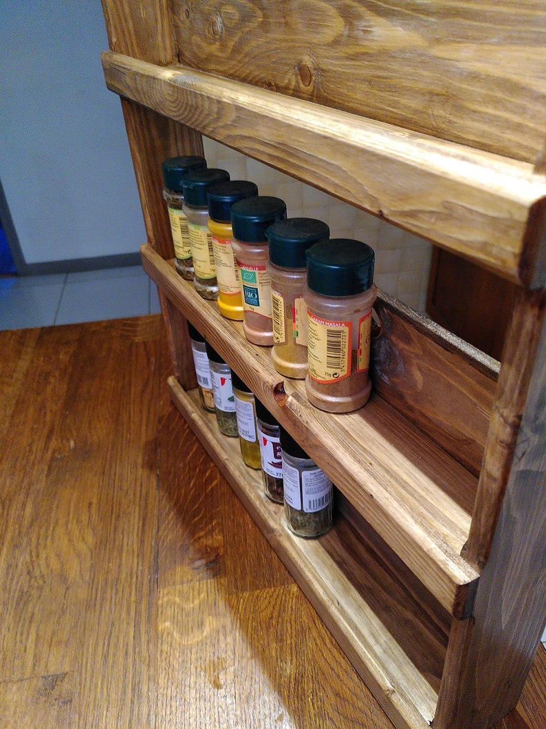 May include: A three-tiered, wooden spice rack with a natural finish. The rack holds several spice jars with black lids, arranged on each shelf. The rack is designed to be mounted on a wall or placed on a countertop for easy access to spices.