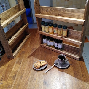 May include: Two wooden spice racks with three shelves each, stained in a warm brown hue. The racks are filled with various spice jars. A plate of cookies and a cup of coffee sit on a wooden table.
