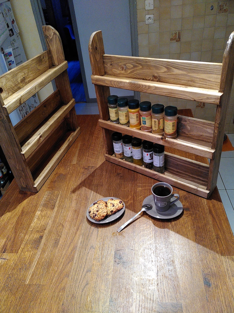 May include: Two wooden spice racks with three shelves each, stained in a warm brown hue. The racks are filled with various spice jars. A plate of cookies and a cup of coffee sit on a wooden table.