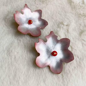 May include: Two flower-shaped earrings. Each earring features a pink and white petal design with a red center. The earrings are set against a soft, white, textured background.