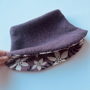 Könnte beinhalten: Ein lilafarbener Fleece-Halswärmer mit einem weißen Blumenmuster als Futter. Der Stoff ist weich und kuschelig.