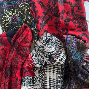 May include: A collection of four folded scarves featuring diverse patterns. One scarf has a black background with colorful paisley designs, another is bright red with black paisley, one is black and white with houndstooth, and one is dark floral.