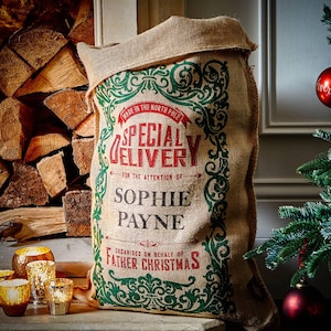May include: A burlap sack with the text "SPECIAL DELIVERY" in red and green, addressed to "SOPHIE PAYNE" and signed by "FATHER CHRISTMAS." The sack is next to a Christmas tree with red ornaments and a stack of firewood.