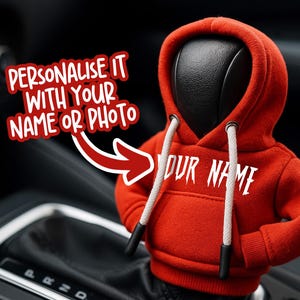 Op de afbeelding: Een rood hoodie-vormig autoaccessoire met de witte tekst "YOUR NAME". De hoodie heeft witte trekkoorden en zwarte uiteinden. De afbeelding bevat ook de tekst "PERSONALIZEER HET MET JE NAAM OF FOTO".