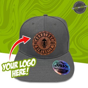 Puede incluir: Una gorra de béisbol gris de visera plana con un parche de cuero marrón con el texto "OVOTONE EST. 2015 NEWCASTLE". La gorra tiene una pegatina en blanco y negro con el texto "OFFICIAL SNAPBACK WEAR Result EST 1990".