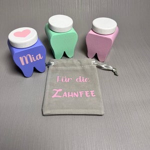 Könnte beinhalten: Drei Holz-Zähne, in Pastellfarben bemalt, mit weißen Deckeln. Der Zahn links ist lila und hat den Namen "Mia" darauf geschrieben. Der mittlere Zahn ist grün und der Zahn rechts ist rosa. Ein kleiner grauer Beutel mit dem Text "Für die ZAHNFEE" in rosa Buchstaben befindet sich vor den Zähnen.