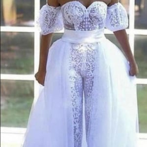 Könnte beinhalten: Ein weißer, schulterfreier Spitzen-Jumpsuit mit einem Tüll-Überrock. Der Jumpsuit hat einen herzförmigen Ausschnitt und kurze Ärmel. Der Rock ist bodenlang und hat eine fließende Silhouette.