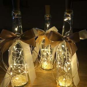 Könnte beinhalten: Drei Glasflaschen mit Korkverschlüssen, gefüllt mit weißen Lichterketten und klaren Perlen. Jede Flasche ist mit einem braunen Satinband verziert, das zu einer Schleife gebunden ist.