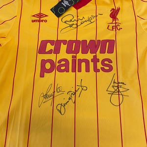 May include: A yellow and red striped soccer jersey with the words "Crown Paints" printed on the front. The jersey has the logo for "Umbro" and "L.F.C." printed on the front. The jersey is signed by several people, including "Kevin Keegan".