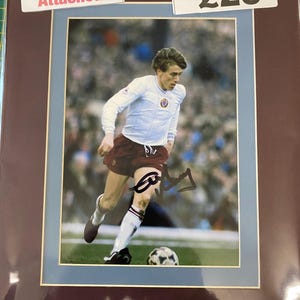 Puede incluir: Una foto enmarcada de un jugador de fútbol con una camiseta blanca con ribetes marrones, regateando el balón. El jugador lleva calcetines blancos y botas negras. La foto está firmada con tinta negra. El marco es marrón con un paspartú azul claro. Una etiqueta blanca con el texto "Photo Proof Attached" está en la esquina superior izquierda. Una etiqueta blanca con el texto "£20" está en la esquina superior derecha.