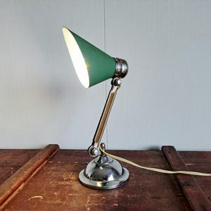 Könnte beinhalten: Eine Schreibtischlampe mit einem konischen grünen Lampenschirm und einem polierten Chrom-Metallsockel mit verstellbarem Gelenkarm. Die Lampe leuchtet und strahlt helles Licht aus dem Inneren des Schirms ab, inklusive weißem Stromkabel.